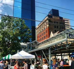A wide shot of people shopping at the Lexington Farmers Market underneath and around the Fifth Third Bank Pavilion. The cityscape can be seen in the background.