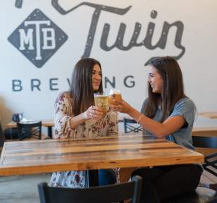 Two women cheers their beers and smile inside the tasting room of Mirror Twin Brewing.