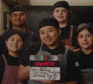 Five student chefs in aprons standing in a commercial kitchen, holding a clapper board with details from the Give Back Gig promo video.