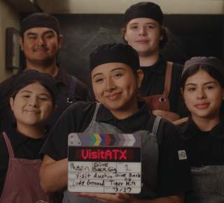 Five student chefs in aprons standing in a commercial kitchen, holding a clapper board with details from the Give Back Gig promo video.