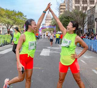 Two women in matching athletic clothes high-fiving in front of the Texas State Capitol Building and finish line of the Austin Marathon.