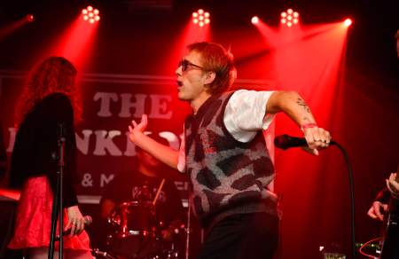 A male singer on a stage wearing sunglasses, holding a microphone, dancing
