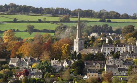 The town of Painswick in the Cotswolds, with it's church steeple rising in the centre, seen from a distance and surrounded by green rolling hills and trees