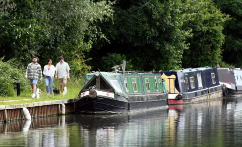 Three friends with a dog walking along the canal towpath past moored canal boats