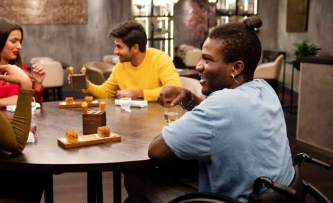 A group of people sit eating, drinking and laughing at a table in a cafe. One of the people is using a wheelchair.