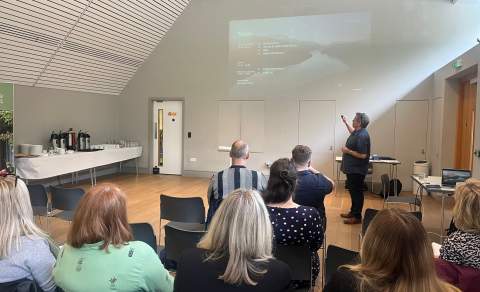 A group of people watch a presentation by a man, who gestures at a slide projection