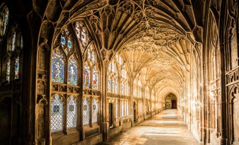 View down the length of the cloisters at Gloucester Cathedral. Sunlight streams through stained glass windows, lighting the detailed fan patterns of the ceiling