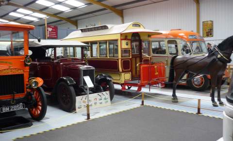 Display at the Oxford Bus Museum featuring a row of historic vehicles, including an early 20th-century motor bus, a red vintage car, a brightly painted horse-drawn carriage, and a classic cream-and-orange coach. A life-sized model horse is harnessed to the carriage, and the exhibit is set within a well-lit indoor space with informational signs and barriers around the displays.