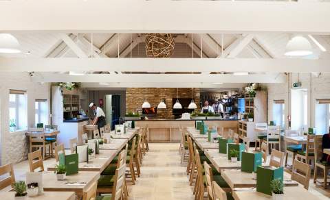 Rows of light wooden dining tables with green menus lead to an open kitchen where chefs are preparing for dinner at Daylesford in the Cotswolds