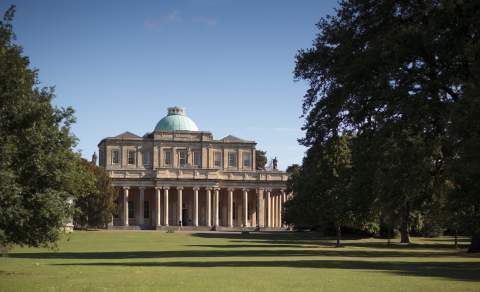 A sweeping lawn with large trees leads to the Grade I listed Regency Pittville Pump Room with green domed roof and large pillars