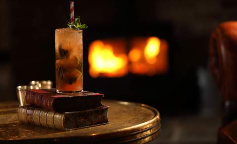 A cocktail in a tall glass with a straw sits on top of two leather-bound books. A fire glows in the background. The Harcourt Arms in the Cotswolds.