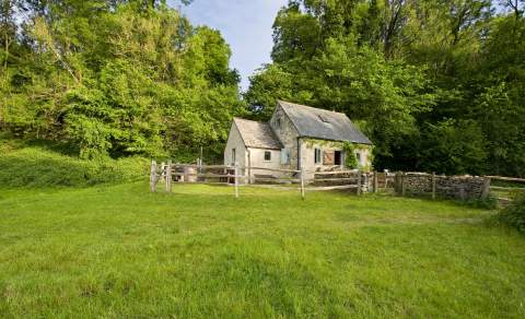 A Cotswold stone cottage surrounded by a wooden fence and backed by trees at Westley Farm