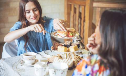 A woman in a blue dress takes a scone from a cake stand, which is filled with sandwiches, scones and cakes, in the Cotswolds