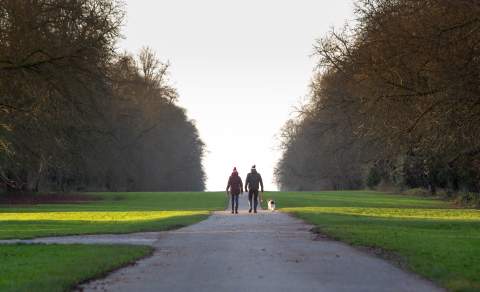 A couple wrapped up warmly and with bobble hats on walking with their dog in Cirencester Park