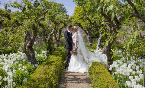 A bride and groom embrace under an archway of trees with white tulip borders at Lords of the Manor