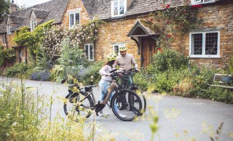 A couple have dismounted their bikes in front of a stunning cottage, with red roses climbing over it's porch, and are looking at their map
