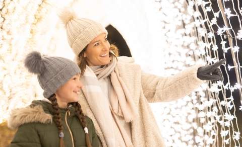 A mother and daughter wearing wooly hats, scarves and gloves in a tunnel of fairy lights