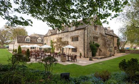 Wooden tables and chairs with white parasols sit on a patio behind The Slaughters Country Inn, surrounded by gardens