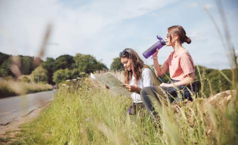 Two women taking a break from walking, one sitting on stone wall drinking from a water bottle, the other reading a map