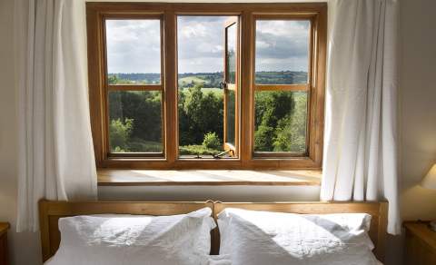 A bed with white linen sits in front of a window framed by crisp white curtains. Through the window you see trees and rolling hills under a cloudy sky. At Heath Farm Holiday Cottages in the Cotswolds
