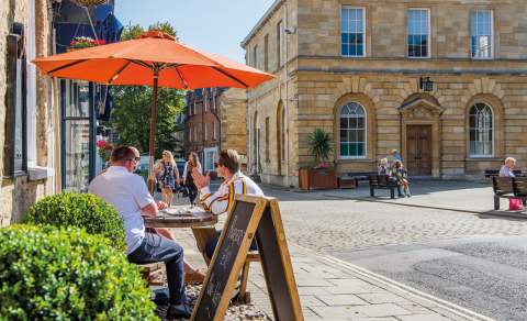 A group of people sit in the shade of an orange parasol outside a cafe, people sit on benches outside the adjacent town hall, whilst two ladies walk along shopping. Woodstock in the Cotswolds