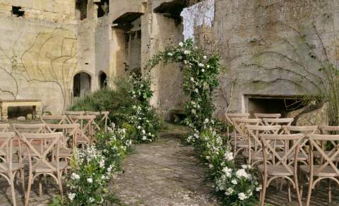 Wooden chairs either side of an aisle decorated with white flowers lead to a white arch of flowers infront of part of Sudeley Castle