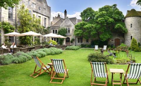 Traditional deckchairs sit on a lawn with flower borders. People sit drinking under sunshades on a patio in front of the 16th century town house at The Close Hotel in the Cotswolds