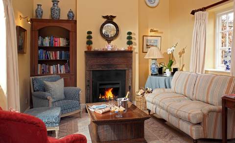 An eclectic mix of sofas and chairs surround a blazing fire with wooden surround in a cozy snug at Bruern Cottages in the Cotswolds