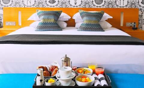 A breakfast tray filled with fresh fruit and pastries rests on a blue ottoman at the foot of a bed at Malmaison Cheltenham