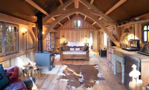 Inside a wooden cabin with large wooden bed, wood burning stove, kitchen, sofa and faux animal skin rug
