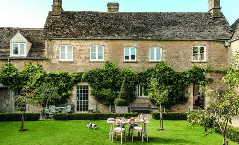 A small dog runs across a lawn dotted with trees in front of a large Cotswold stone cottage covered in wisteria at Daylesford Cottages in the Cotswolds