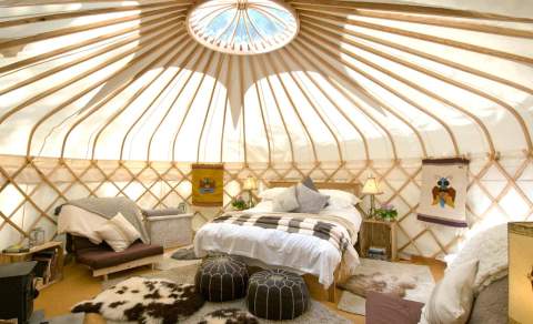 A circular skylight illuminates a cozy double bed, faux animal skin rugs and pouffes dotted in front of a wood burner inside Sapperton Yurt, at Westley Farm in the Cotswolds