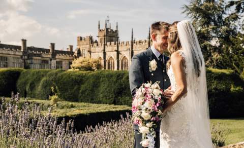 A bride and groom stand face-to-face, smiling, in a garden with Sudeley Castle in the background