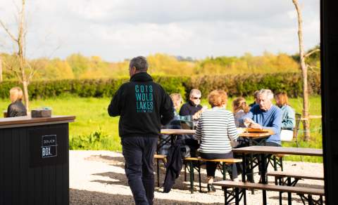 A staff member takes drinks to a group of customers sat in the outdoor seating at Cotswold Lakes Brew Co in the Cotswolds