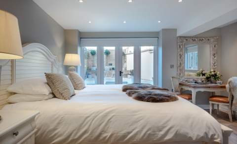 A king-size bed with crisp white bedding stands in a neutrally decorated room with doors leading to the garden at Woolmarket House in the Cotswolds