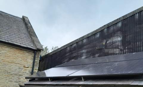 Modern building roof fitted with a row of solar panels, set beside a stone structure under an overcast sky.