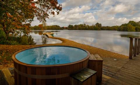 A wooden hot tub on a deck with views across the lake and leaves falling from the trees, at Log House Holidays in the Cotswolds