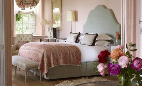 A vase of pink peonies sits on a table in front of a view of a large bedroom with upholstered bed, pale pink walls and large sash window at Thyme in the Cotswolds