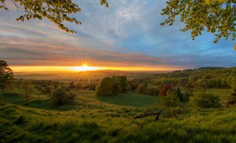 The sun rises over the horizon, illuminating the sky with a warm yellow glow over the lush green fields and trees surrounding the Cotswold Way