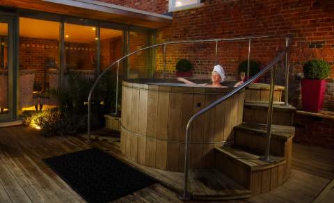 A lady relaxes at night in an outdoor wooden hot tub, surrounded by plants and subdued lighting at The Greenway Hotel & Spa in the Cotswolds