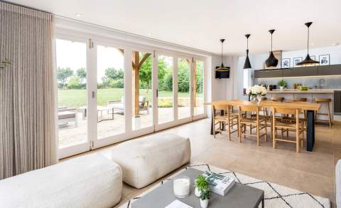 An open plan living space with modern kitchen, wooden dining table and elegant seating area, opening out through bi-fold doors to a patio. StayCotswold in the Cotswolds