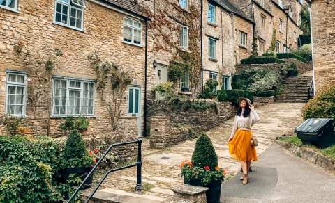 A woman wearing a hat smiles as she poses in front of stone cottages on a stepped hill in Tetbury