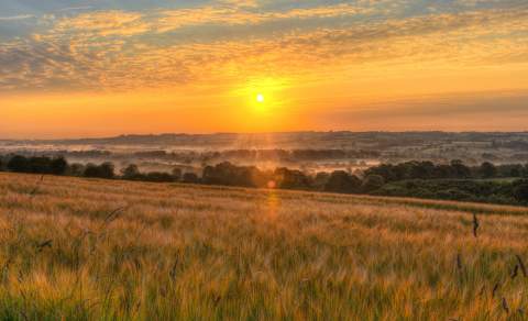 The sun shines warm yellow through wispy clouds over open countryside, where mist lingers in the valleys