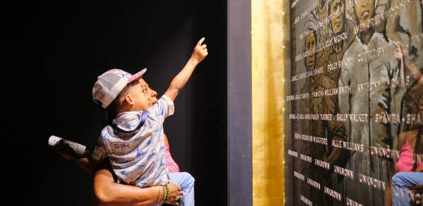 African American man holding a young child in front of the plaque with engraved names of the Clotilda, the Last American Slave Ship
