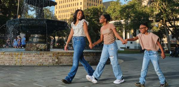 Two Black girls and one Black boy walk in a park past a fountain with buildings in the background