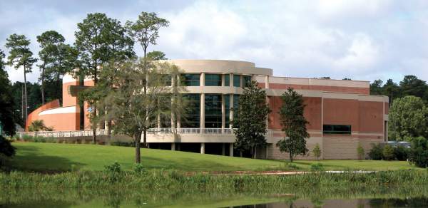 The Mobile Museum of Art building with curved architecture reflects in a serene pond, surrounded by lush green grass and trees.