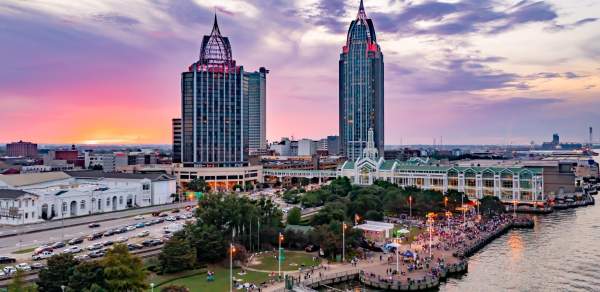 A vibrant sunset over Downtown Mobile skyline, featuring tall buildings, a bustling waterfront, and a lively crowd below.