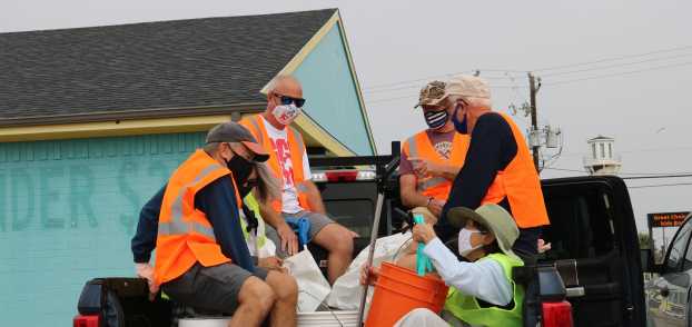 A group of five volunteers in orange safety vests sit in the bed of a truck with trash bags and buckets