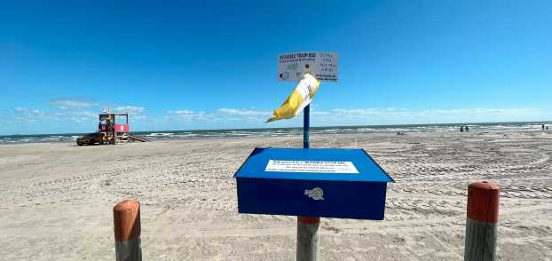 In the foreground is a blue metal box mounted on a wooden pole. From it hangs a yellow mesh bag. In the background is the beach and a lifeguard station.