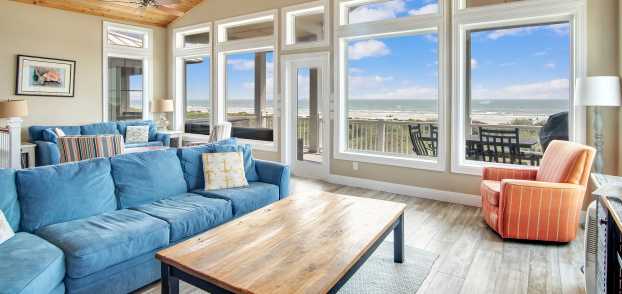 Photo of a living room interior with ceiling to floor glass windows looking out to the beach. An orange chair, blue couches, lamps, and coffee tables are visible.